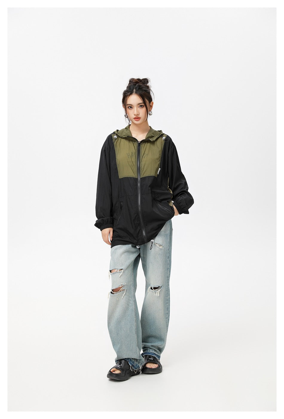 Model in black and olive color-block hooded jacket, hand in pocket, with ripped light-wash jeans and black sandals