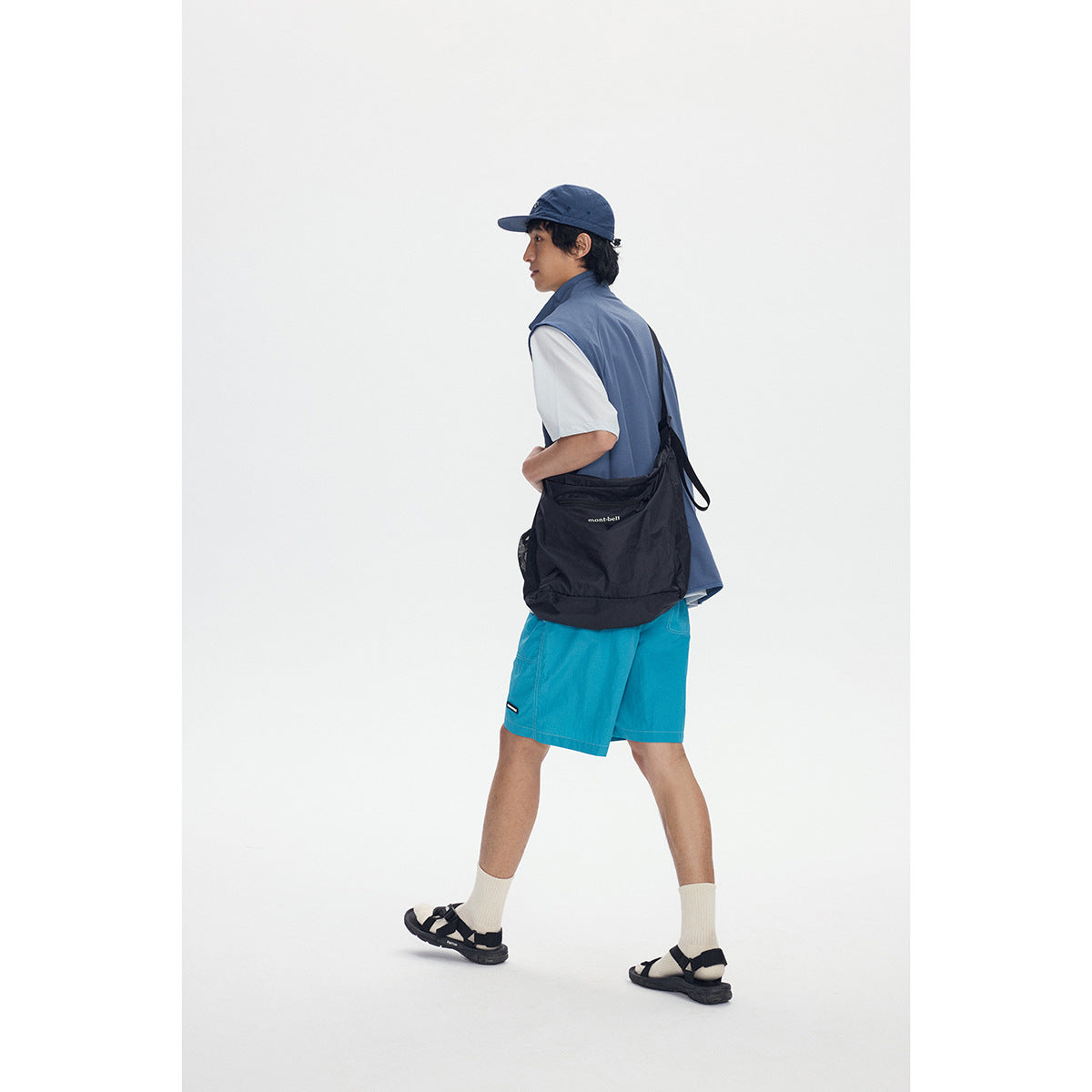 Rear three-quarter view of a model wearing a slate-blue vest, white T-shirt, turquoise shorts, and black shoulder bag.