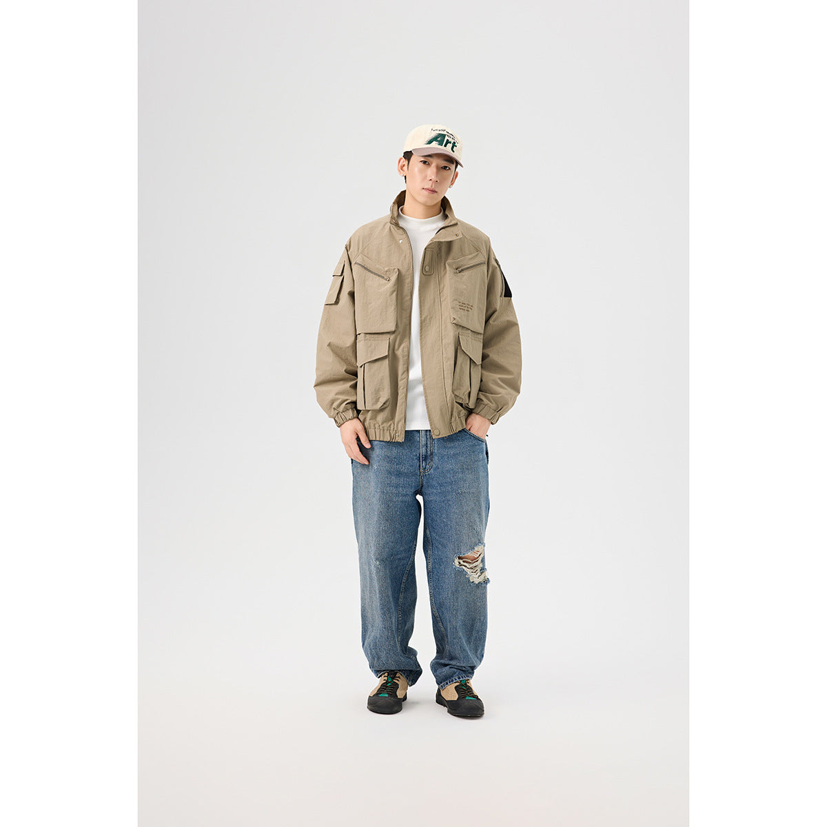 Full-length model wearing a khaki utility jacket with multiple flap and zip pockets, blue jeans, and a cream cap.