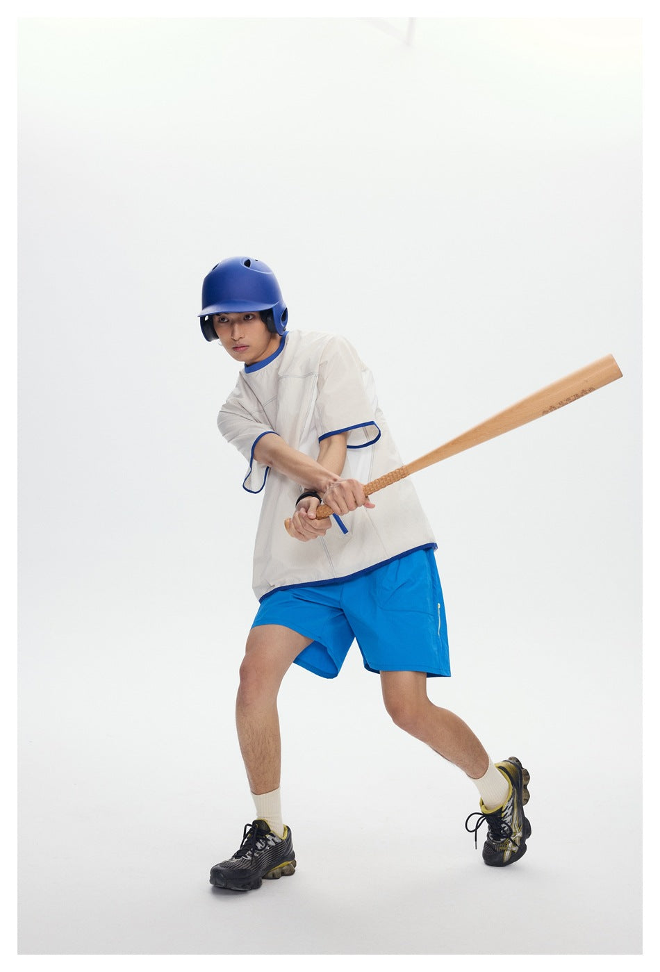 Model swings a bat while wearing an oversized cream shirt with blue trim and bright blue shorts.