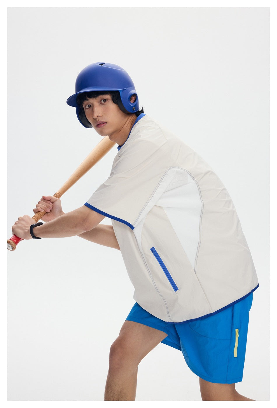 Model in a cream shirt with blue trim and bright blue shorts, posing with a bat in a blue helmet.