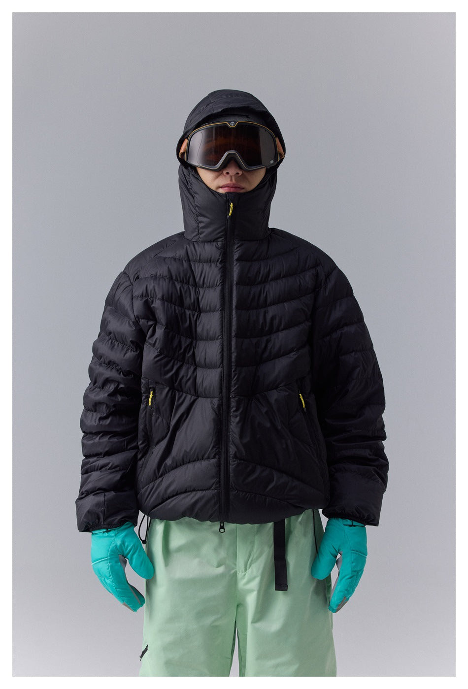 Front view of a model wearing a black quilted hooded jacket, ski goggles, and turquoise gloves.