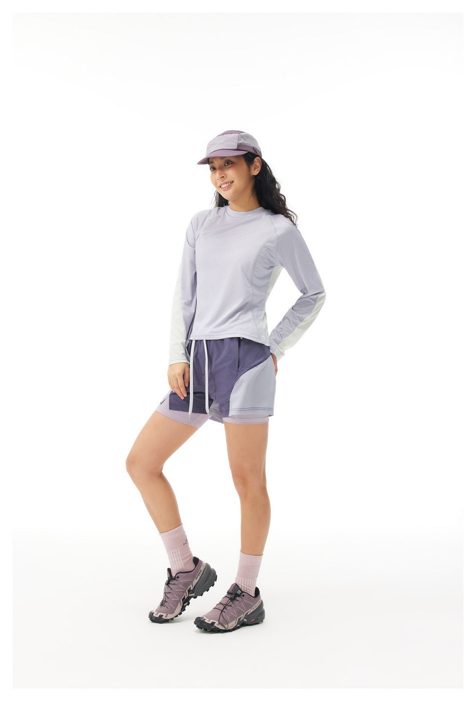 Woman in a lavender long-sleeve top with off-white sleeve panels, shorts and a cap, three-quarter view