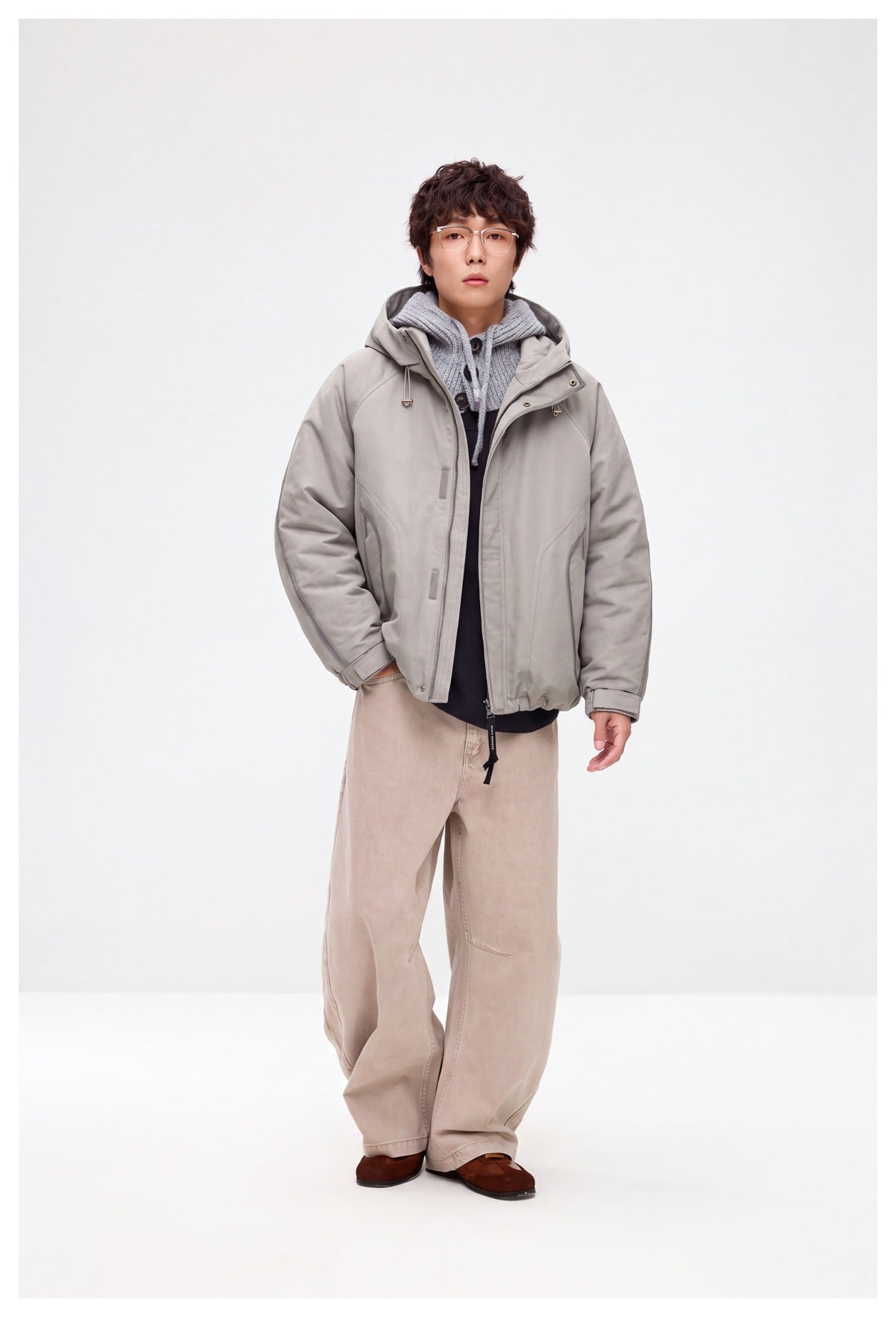 Full-length view of a model wearing an open light gray hooded jacket with layered knitwear and loose beige trousers.