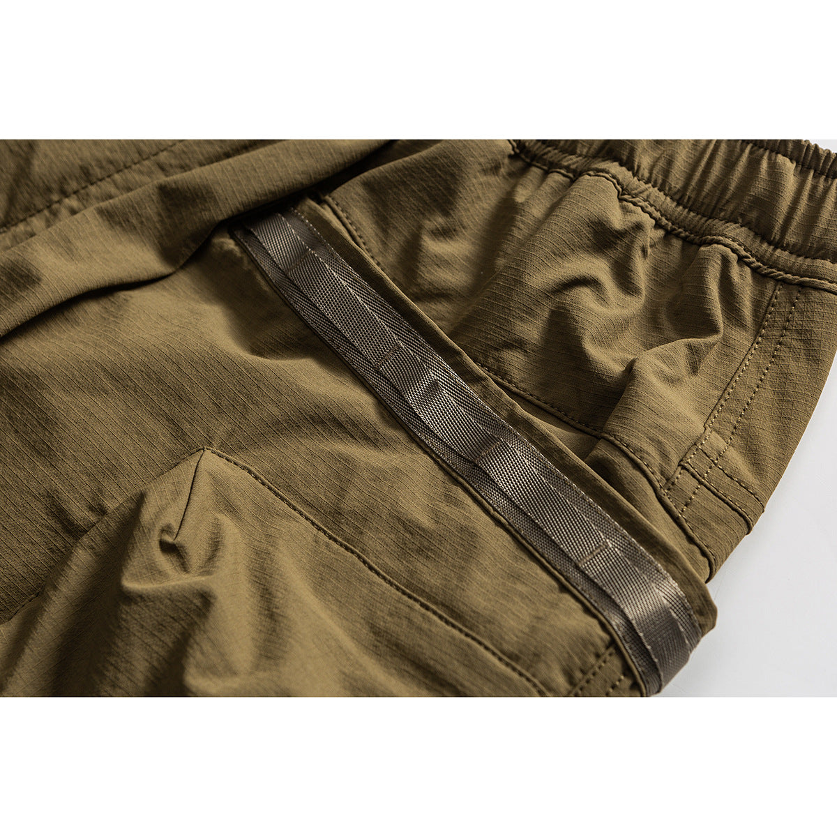 Close-up of olive cargo shorts showing a webbing belt, elastic waistband, stitching, and side pocket.