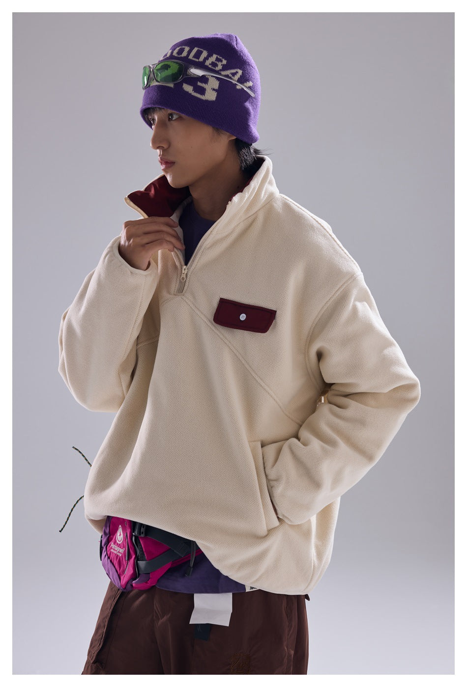 Model wearing cream half-zip fleece-like jacket with maroon chest pocket flap, with beanie and brown pants