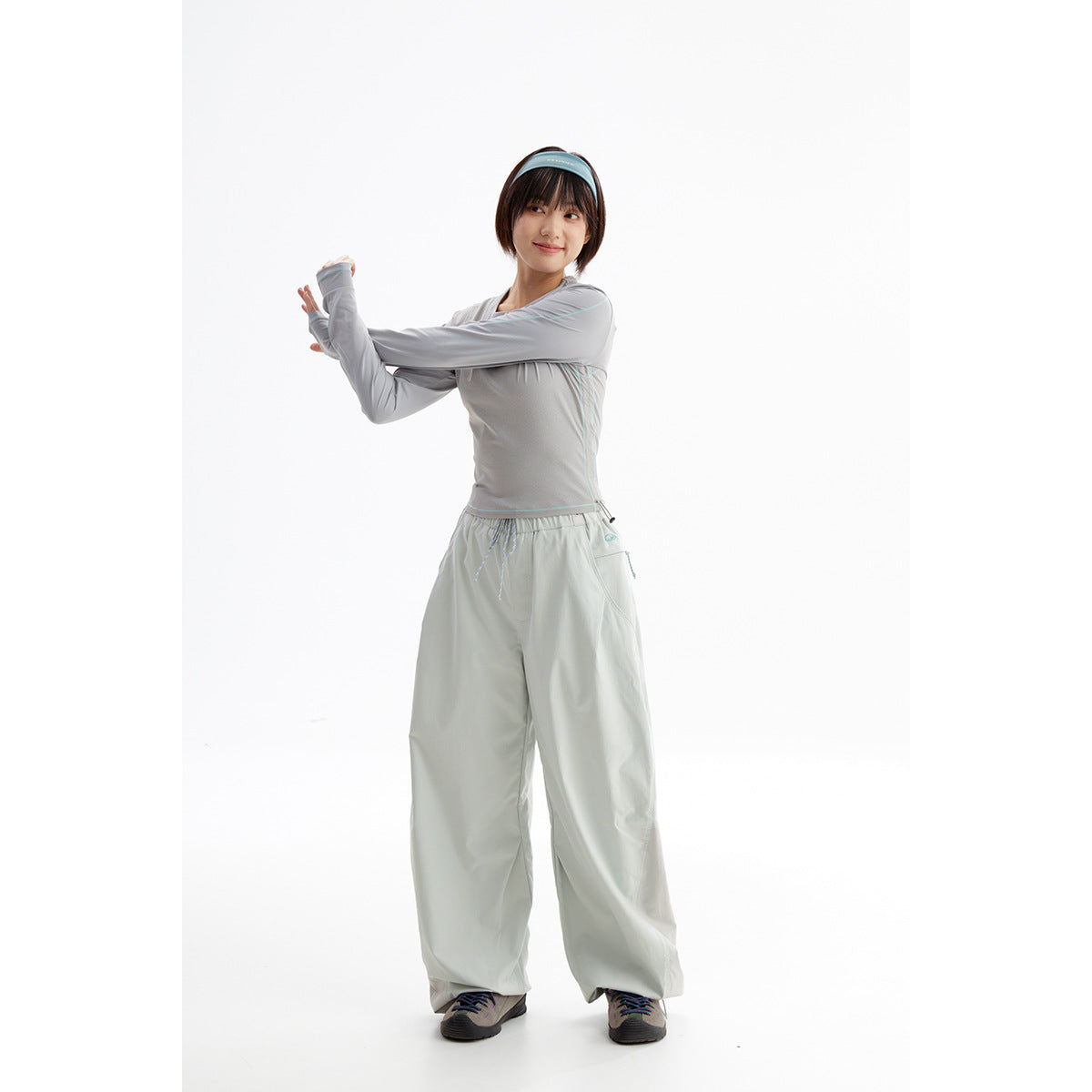 Model stretching in a light gray long-sleeve top with turquoise piping and pale gray wide-leg pants.