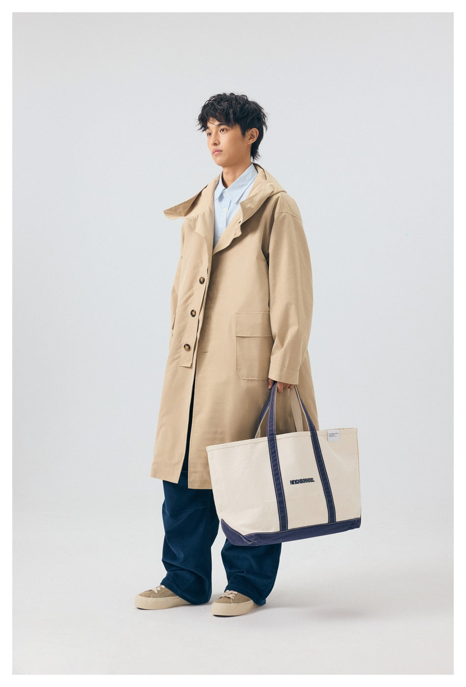 Side view of a model in a khaki long hooded coat with a high collar and flap pocket, holding a tote