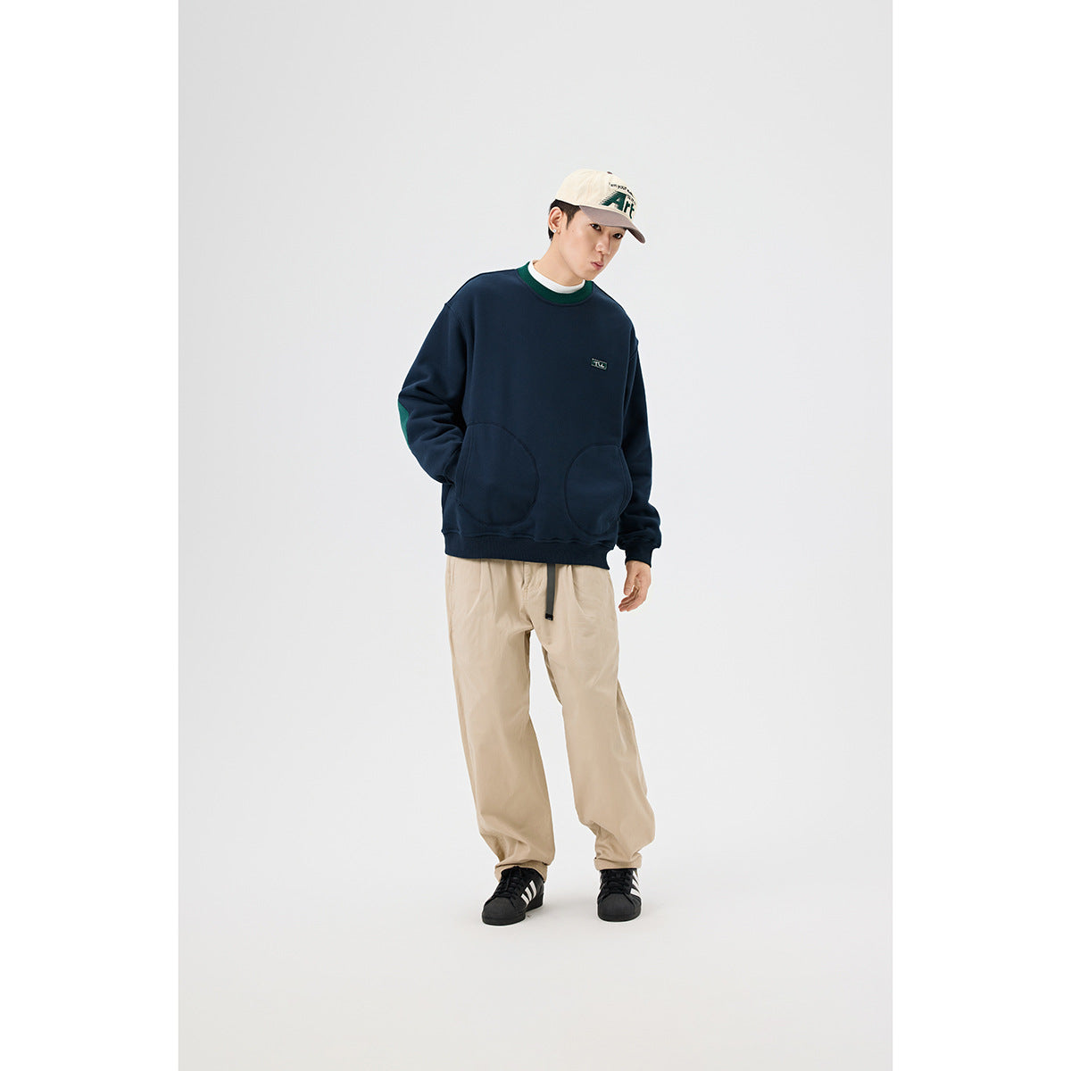Model wearing a navy crewneck sweatshirt with green trim and two rounded front pockets, beige trousers, and a cream cap.
