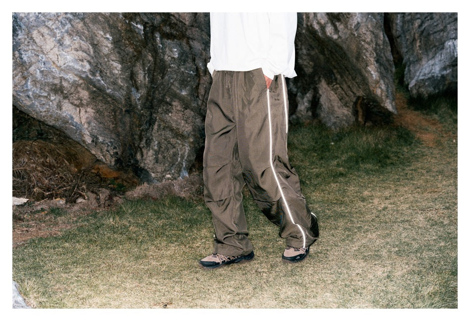 Angled view of loose taupe-gray pants with reflective side piping, worn by a model outdoors.