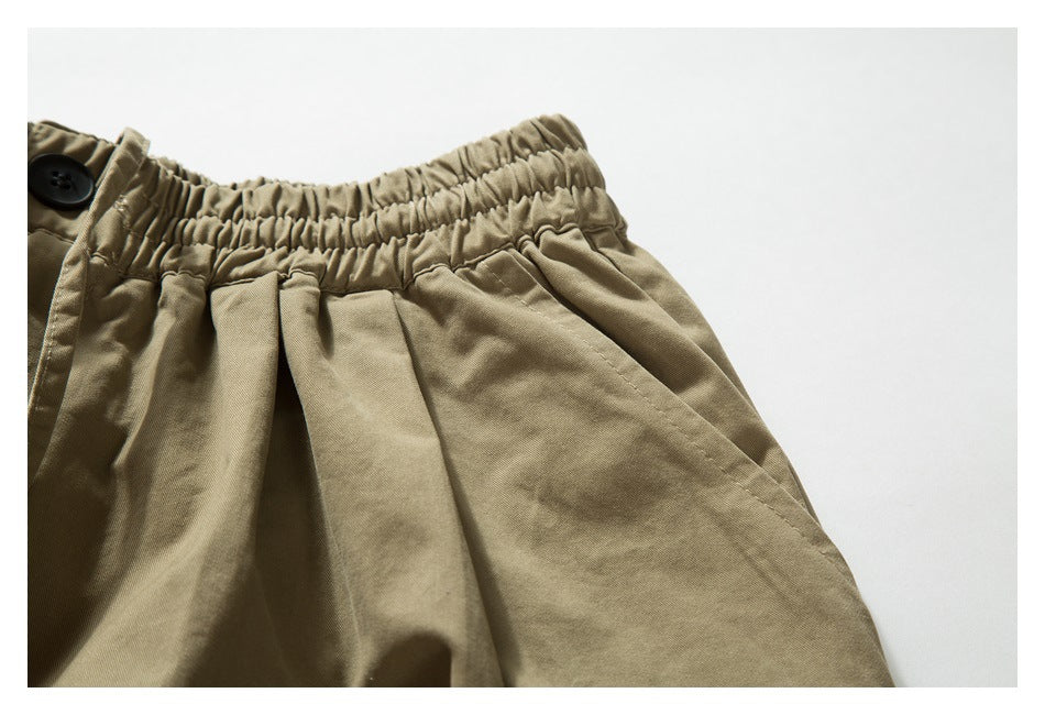 Close-up of khaki trousers showing the gathered elastic waistband, button, pleats, and pocket opening.