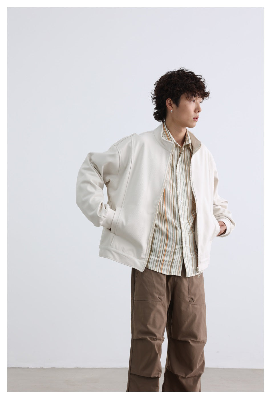 Model wearing an open off-white zip jacket over a striped shirt and taupe trousers, turned to the side.