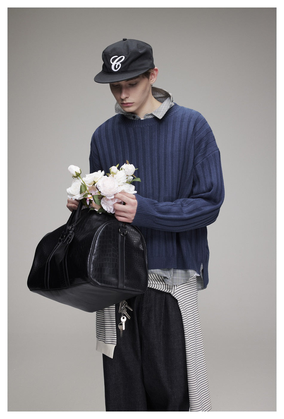 Closer view of a model wearing a blue ribbed sweater over a checked shirt, with a black cap and dark jeans.
