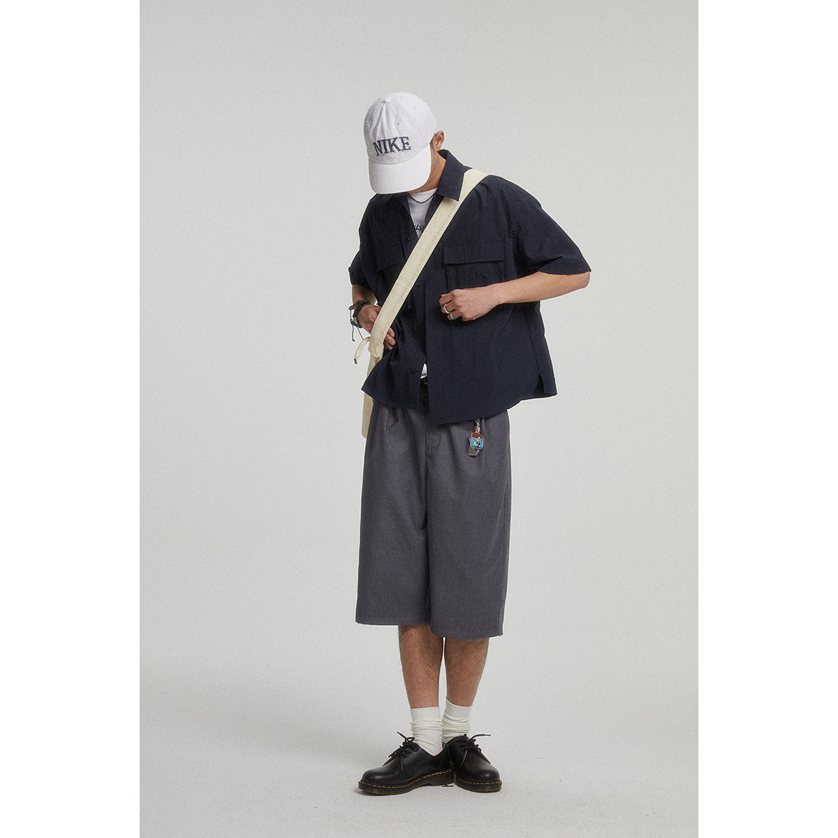 Model wearing a navy short-sleeve overshirt with chest pockets and loose charcoal shorts, styled with a white cap and cream