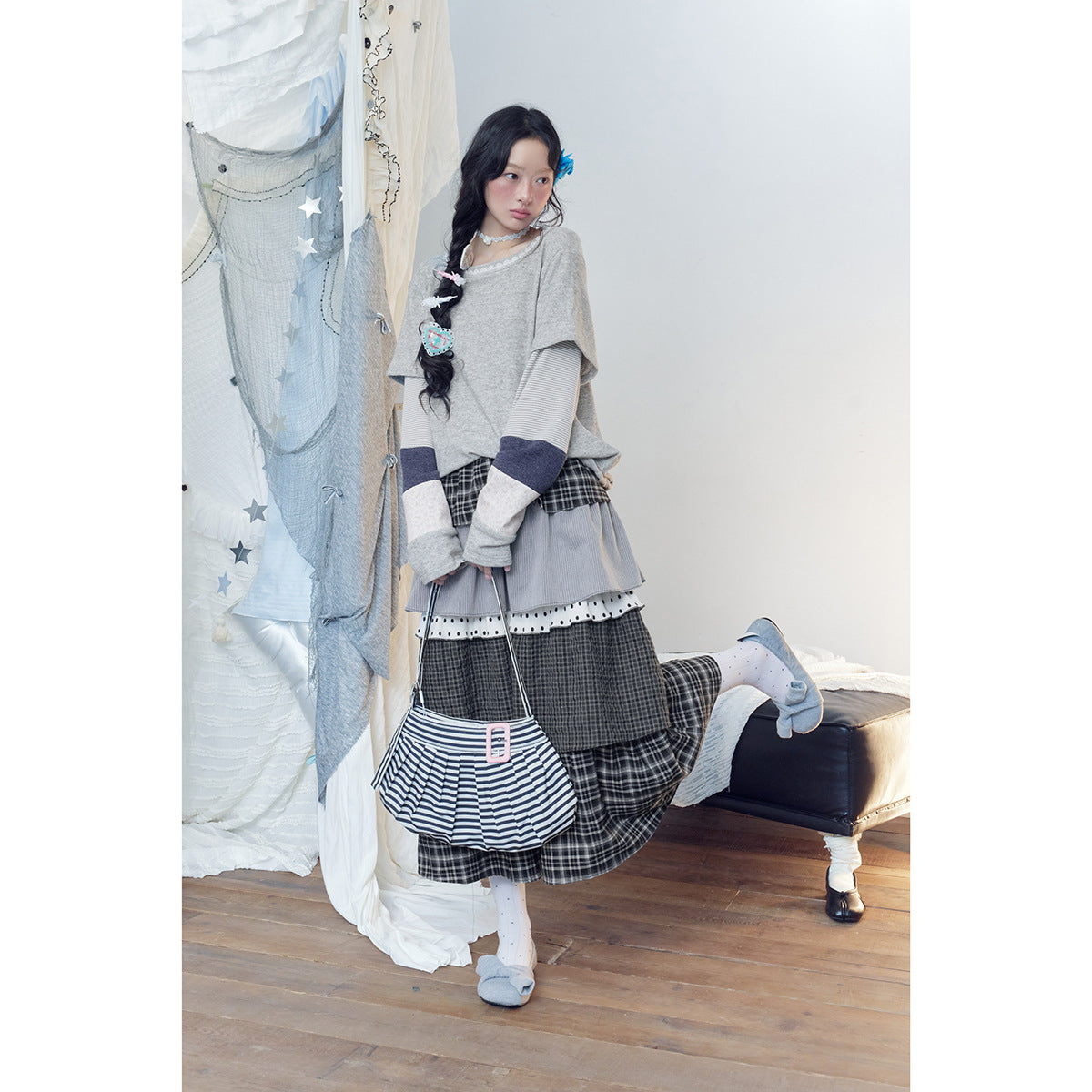 Model posing in a gray patchwork-sleeve top and layered dark plaid skirt, holding a striped bag.