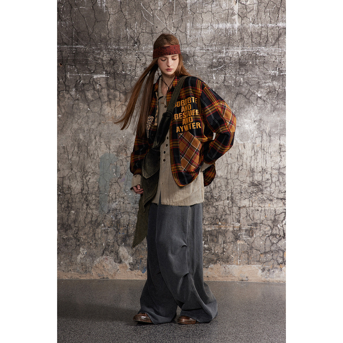 Full-length model wearing a black, red, and mustard plaid patchwork shirt over a beige top and wide charcoal trousers.