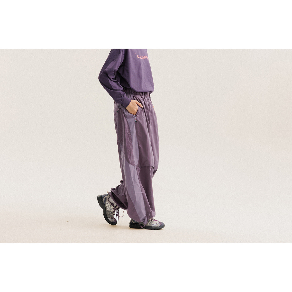 Side view of a model wearing loose purple pants with an elastic waist and gray trail shoes.