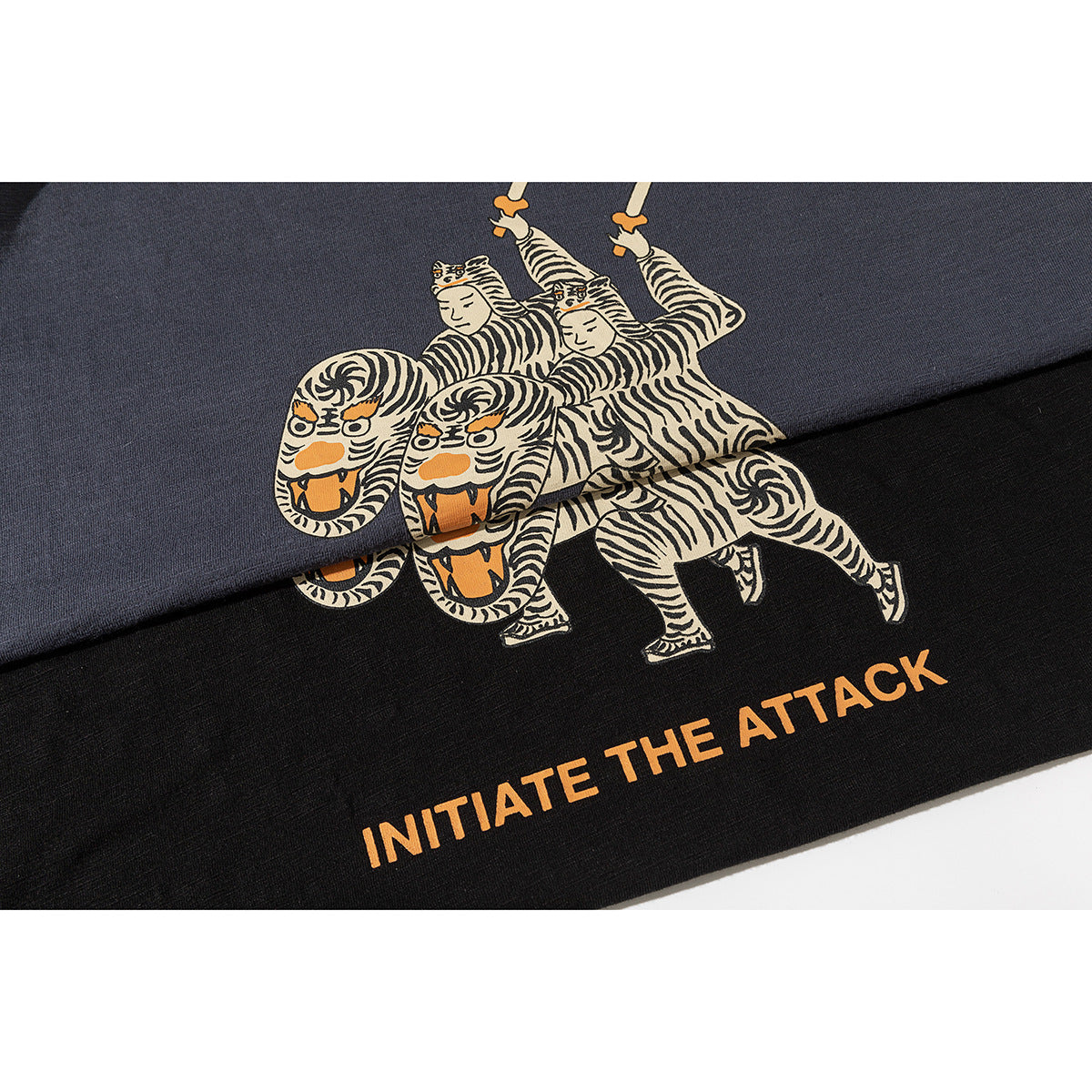 Close-up of cream-and-orange tiger-rider artwork and “INITIATE THE ATTACK” text printed on charcoal and black T-shirts.