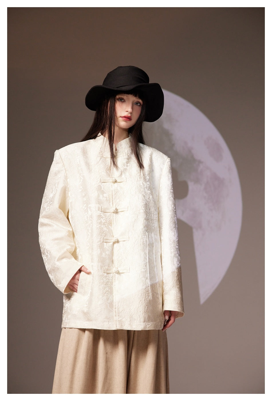 Model wearing an ivory floral-textured jacket with frog closures, a black wide-brim hat, and a beige skirt.