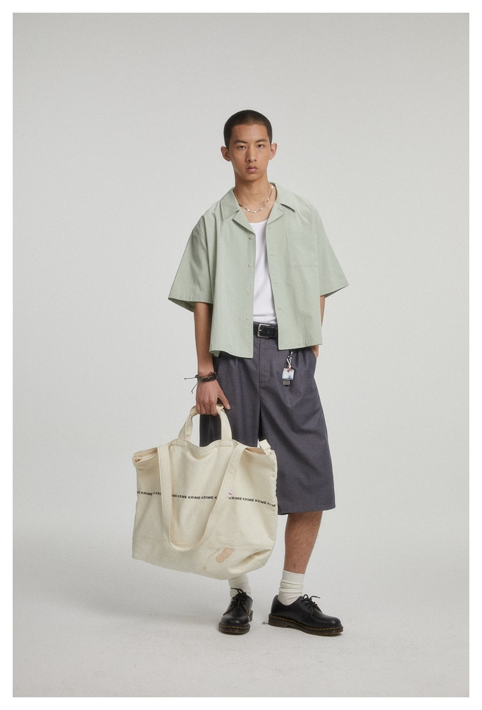 Model wearing an open pale sage short-sleeve shirt with a chest pocket and charcoal shorts.