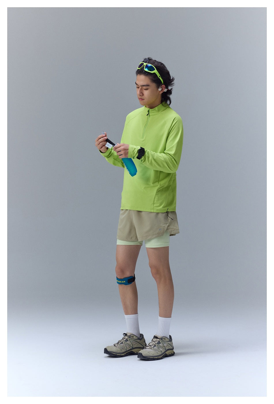 Person in lime green quarter-zip top with beige shorts holding a blue soft flask, angled side view
