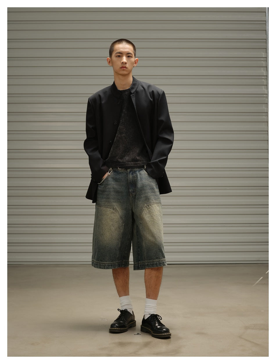 Model wearing black stand-collar jacket open over a dark tee with faded denim shorts and black shoes