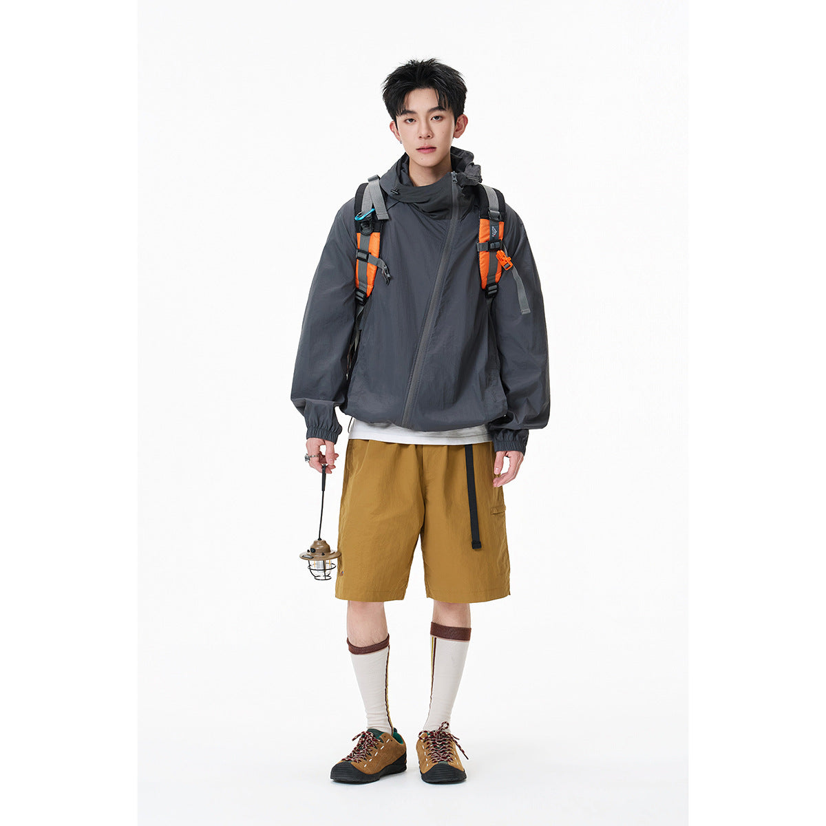 Model wearing a charcoal hooded windbreaker with mustard shorts and an orange-accented backpack.