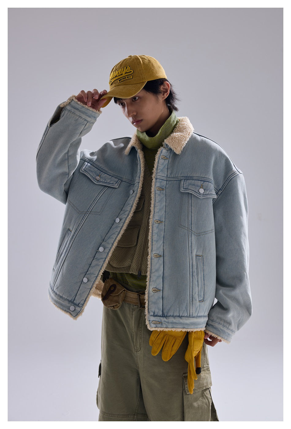 Model wearing light blue denim jacket with fleece collar, open over a green top, touching a mustard cap