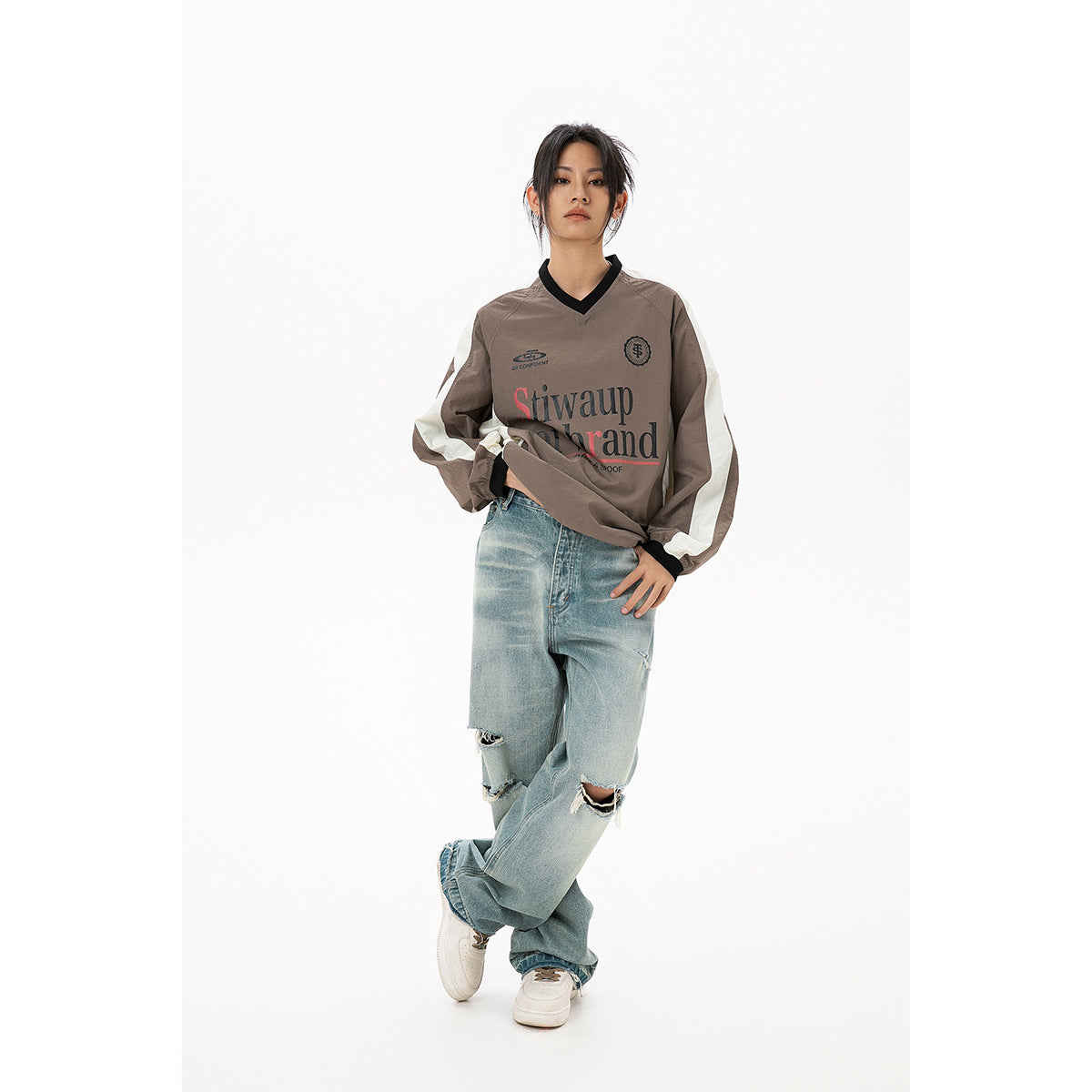 Model wearing a taupe and cream V-neck pullover with black trim and large front lettering.