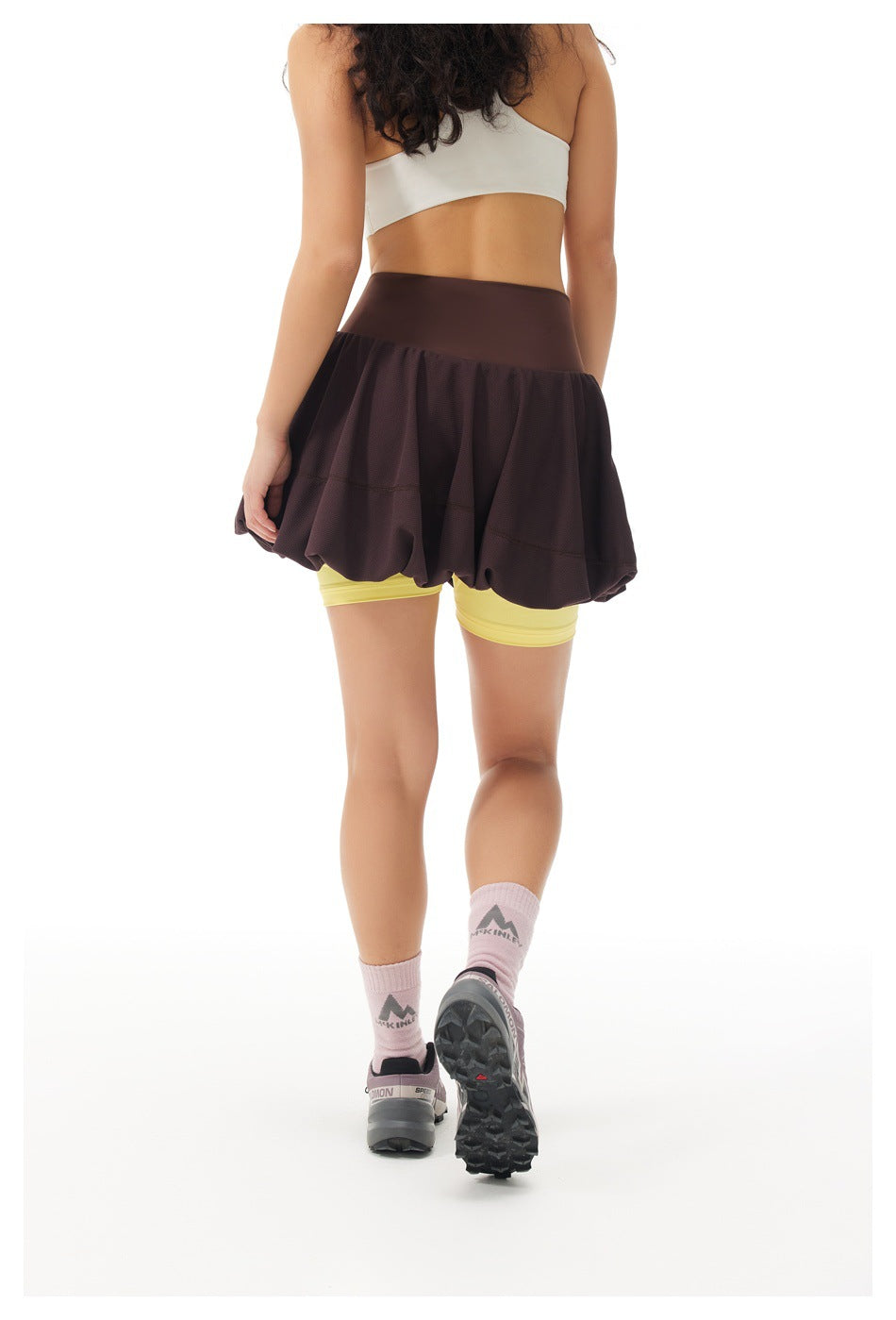 Back view of woman wearing brown bubble-hem skort with yellow inner shorts, white top, pink socks and sneakers