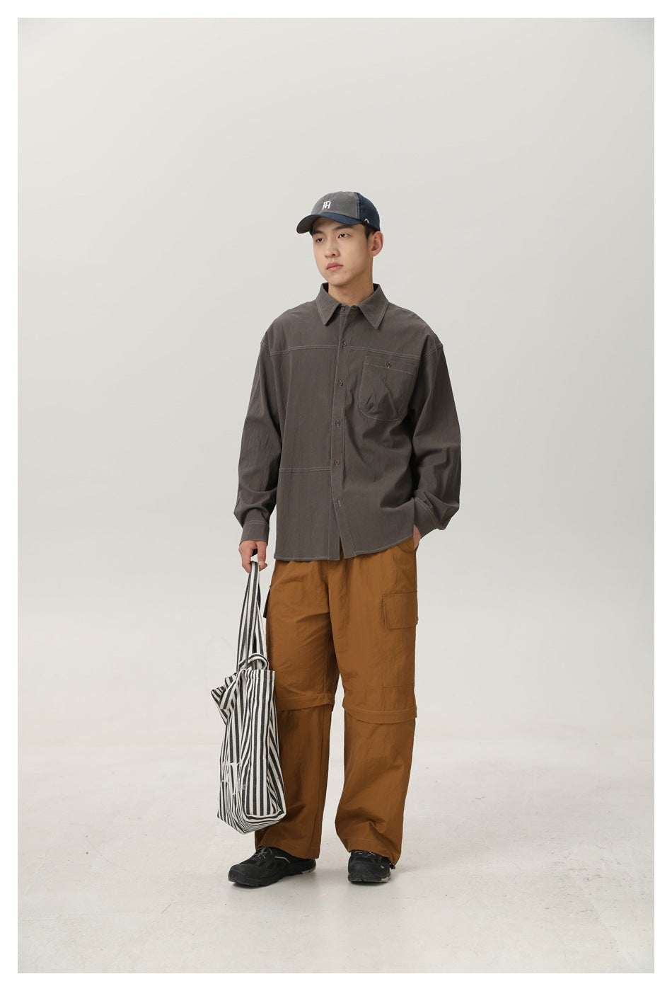 Full-length front view of a man wearing a charcoal gray button-up shirt and brown cargo pants, holding a striped tote.