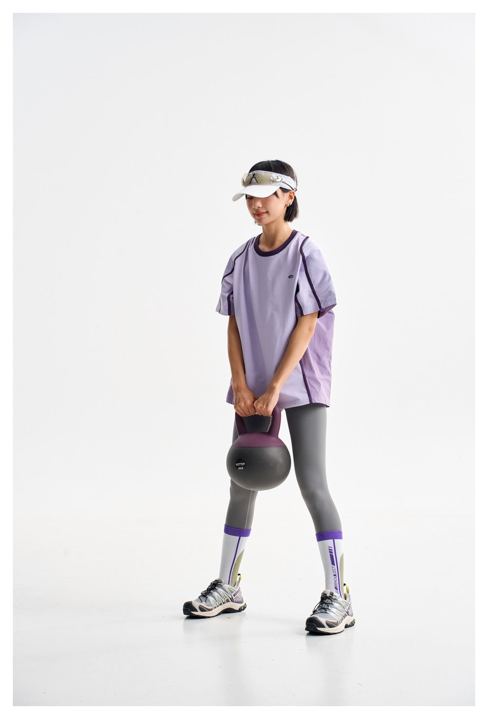 Model in a lavender oversized T-shirt and charcoal leggings, holding a kettlebell in a studio.