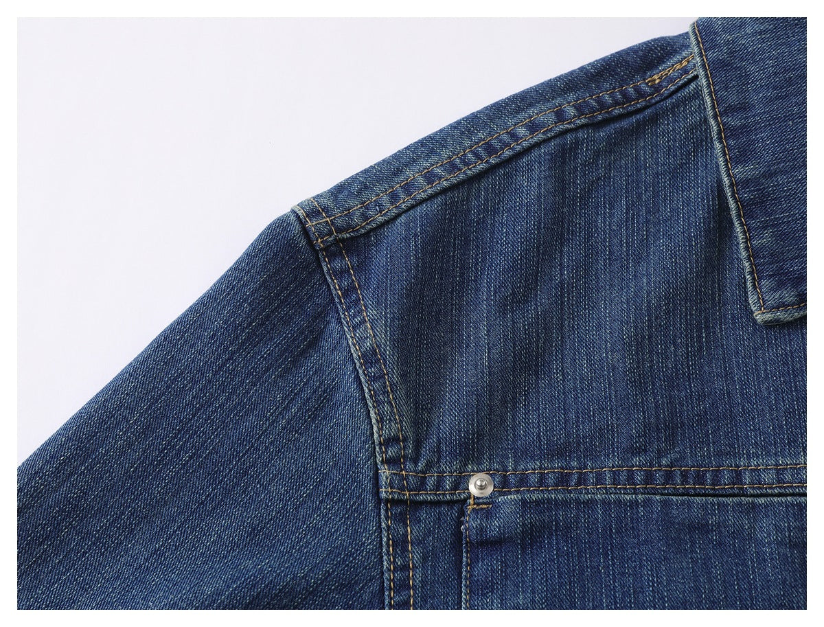 Close-up of a dark blue denim jacket shoulder and chest seam with contrasting tan stitching and a metal rivet.