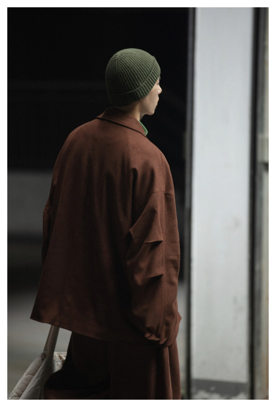 Rear view of a model in a brown coat and olive ribbed beanie, carrying a light-colored bag.