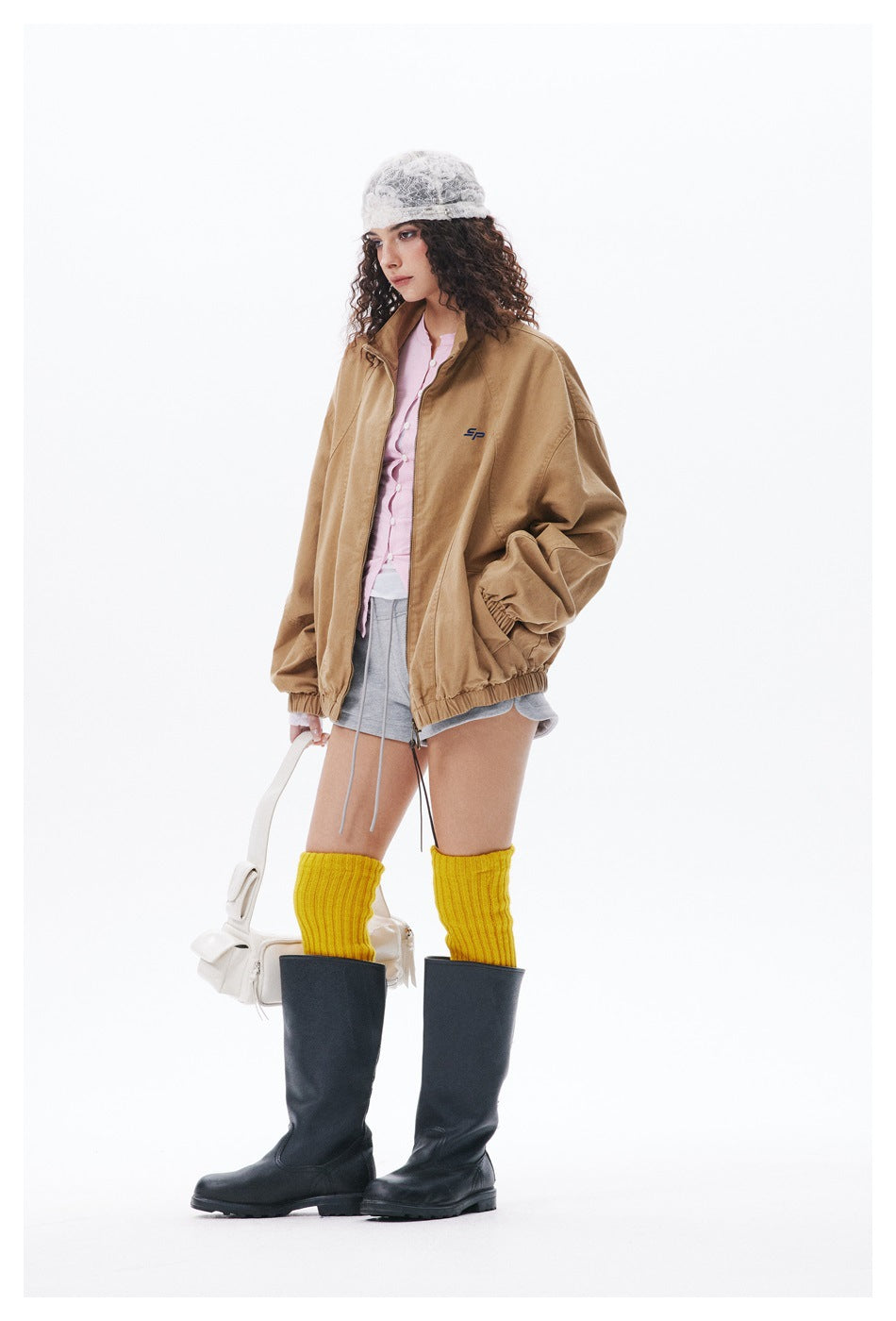 Model wearing the khaki zip jacket open over a pink top, gray shorts, yellow knee socks and black boots, with a hand in the