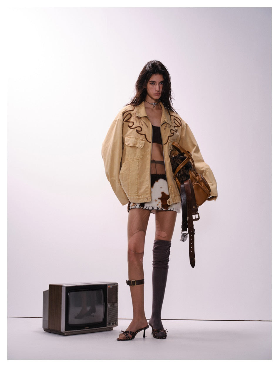 Model in an oversized sand-brown embroidered western jacket with a tan bag, cow-print skirt and a vintage TV beside her