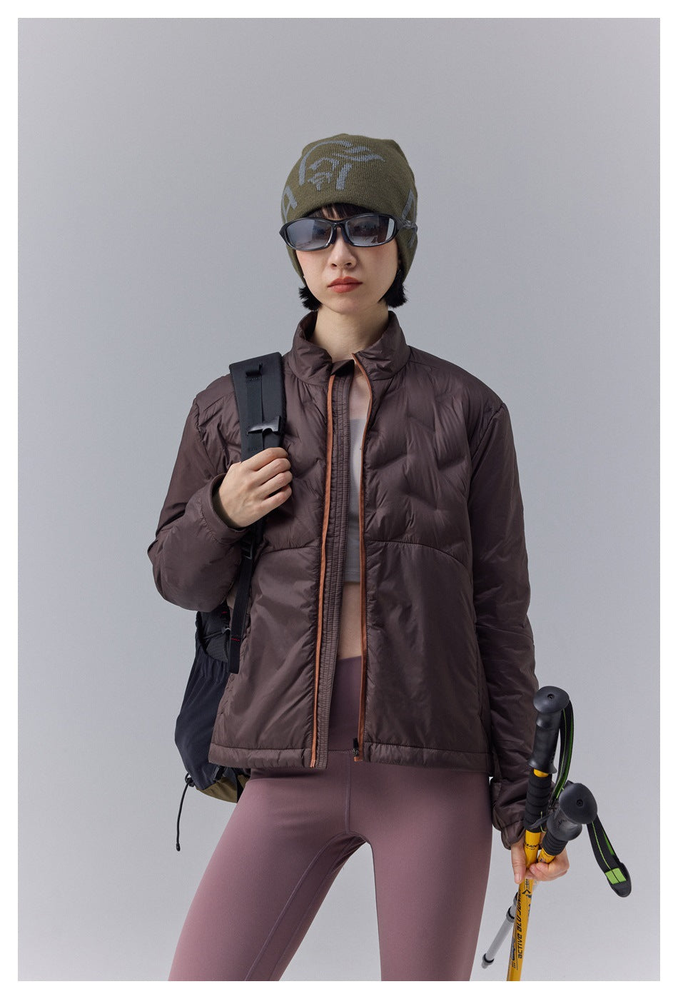Front view of a model in a brown zip-front puffer jacket and mauve leggings, wearing an olive beanie and sunglasses.