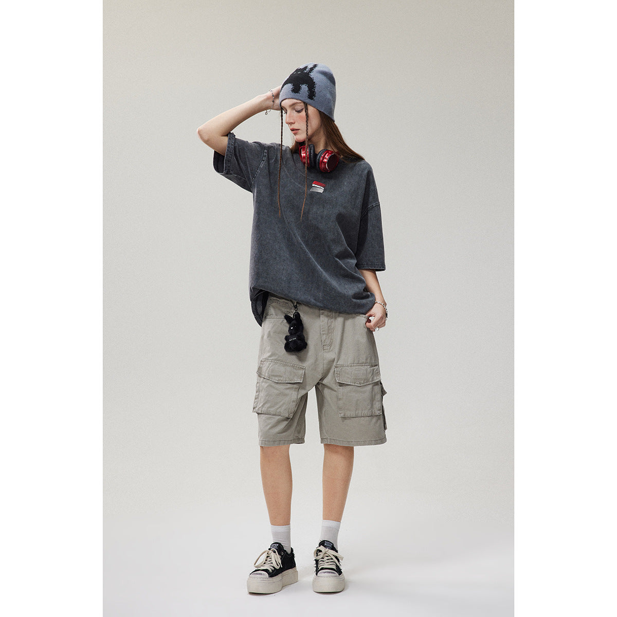 Model wearing an oversized charcoal gray T-shirt with a small chest graphic, khaki cargo shorts, and a gray patterned beanie.