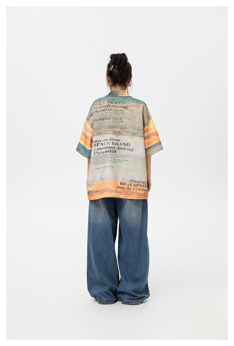 Back view of a model wearing a sunset-print short-sleeve shirt with large slogan text and wide-leg blue jeans