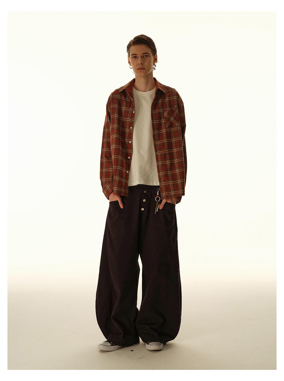 Full-length model wearing an open rust plaid shirt over a cream tee and loose, wide-leg black trousers.