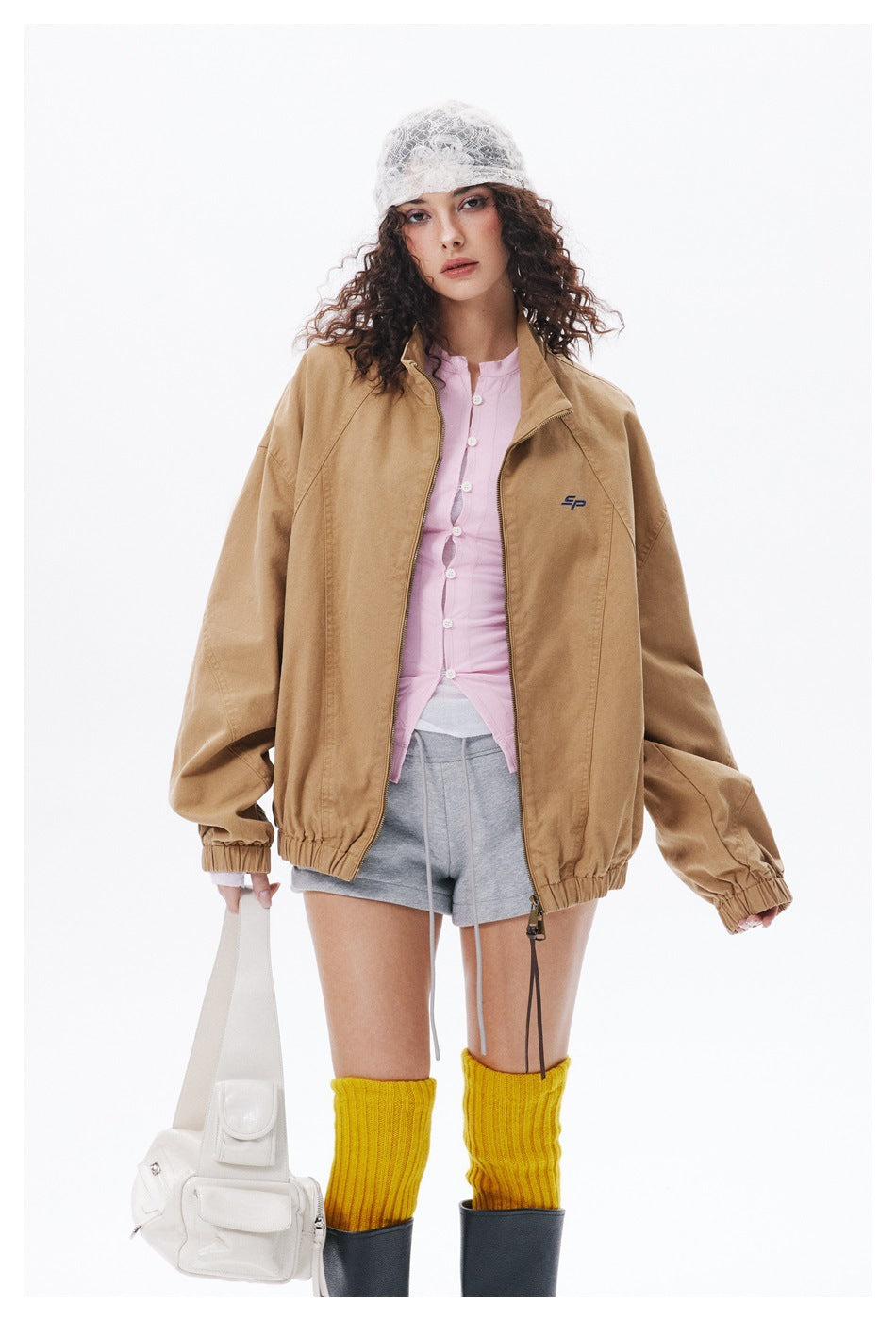 Model wearing the oversized khaki zip jacket open over a pink buttoned top and gray shorts, holding a white bag