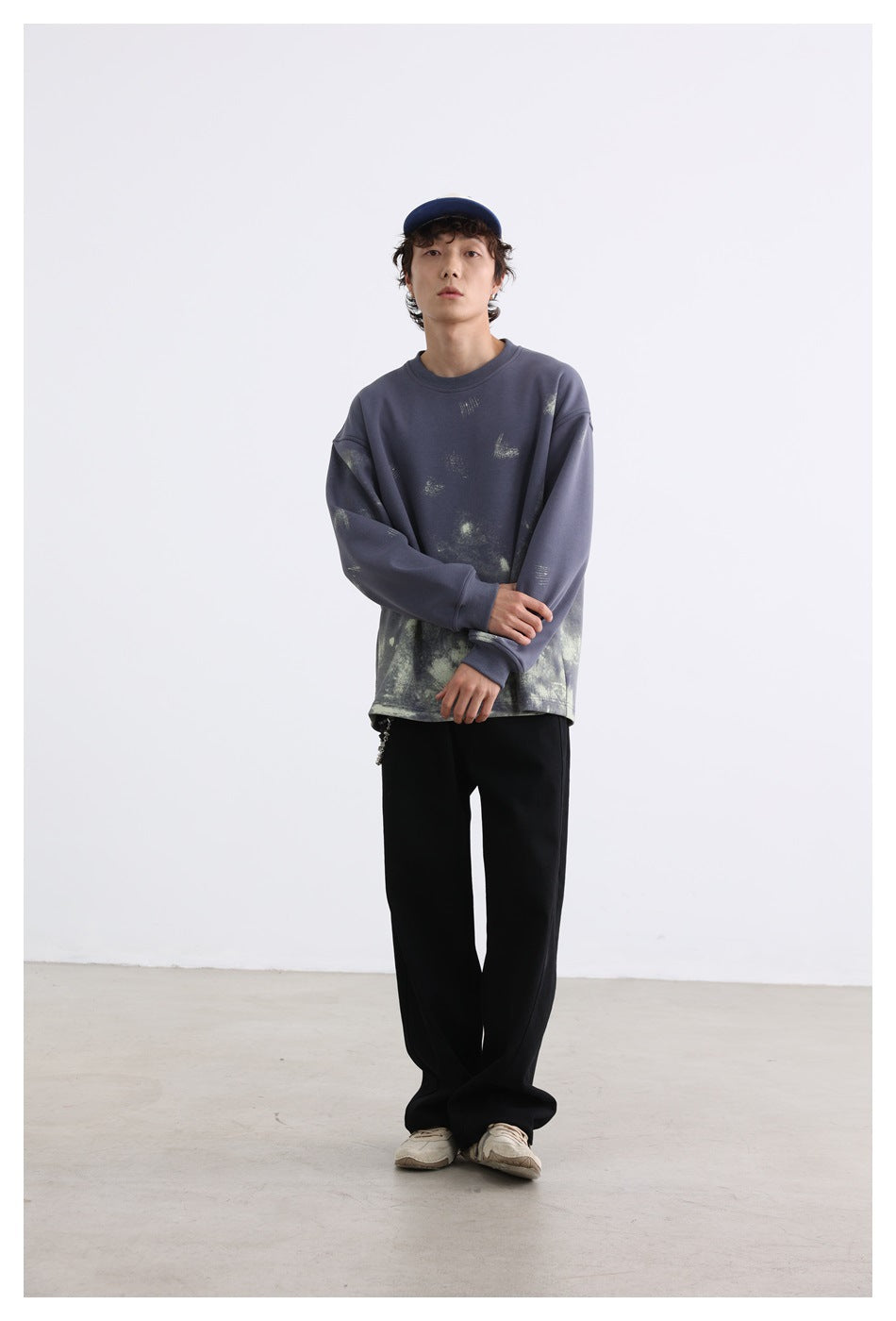 Model wearing an oversized slate-blue sweatshirt with pale distressed markings, black wide-leg trousers, and a navy cap.