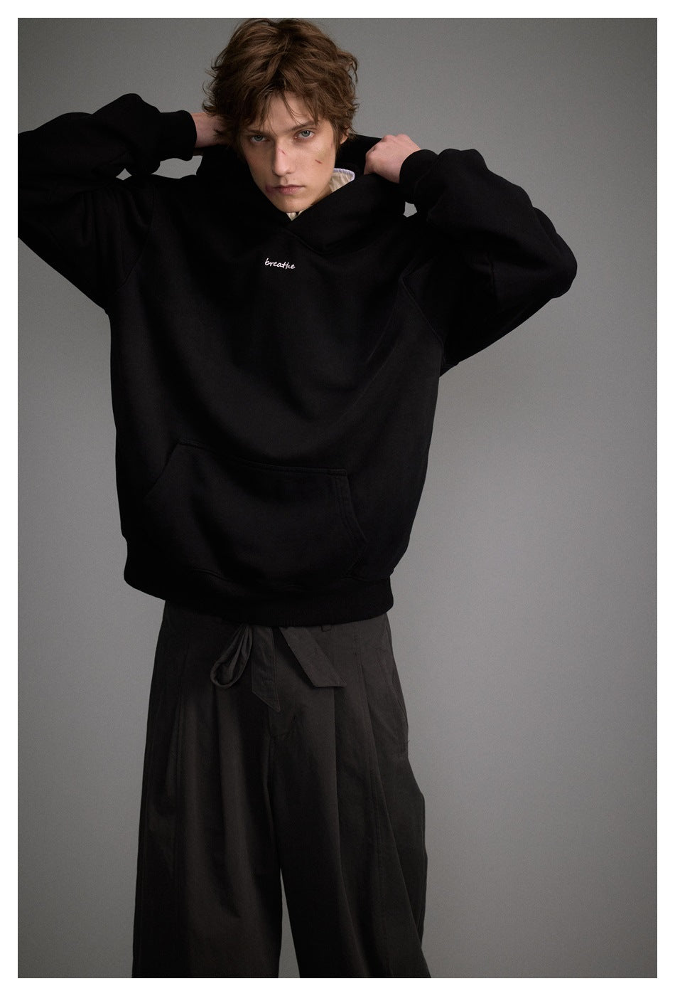 Model adjusting the hood of a black oversized hoodie with a kangaroo pocket and white chest script.