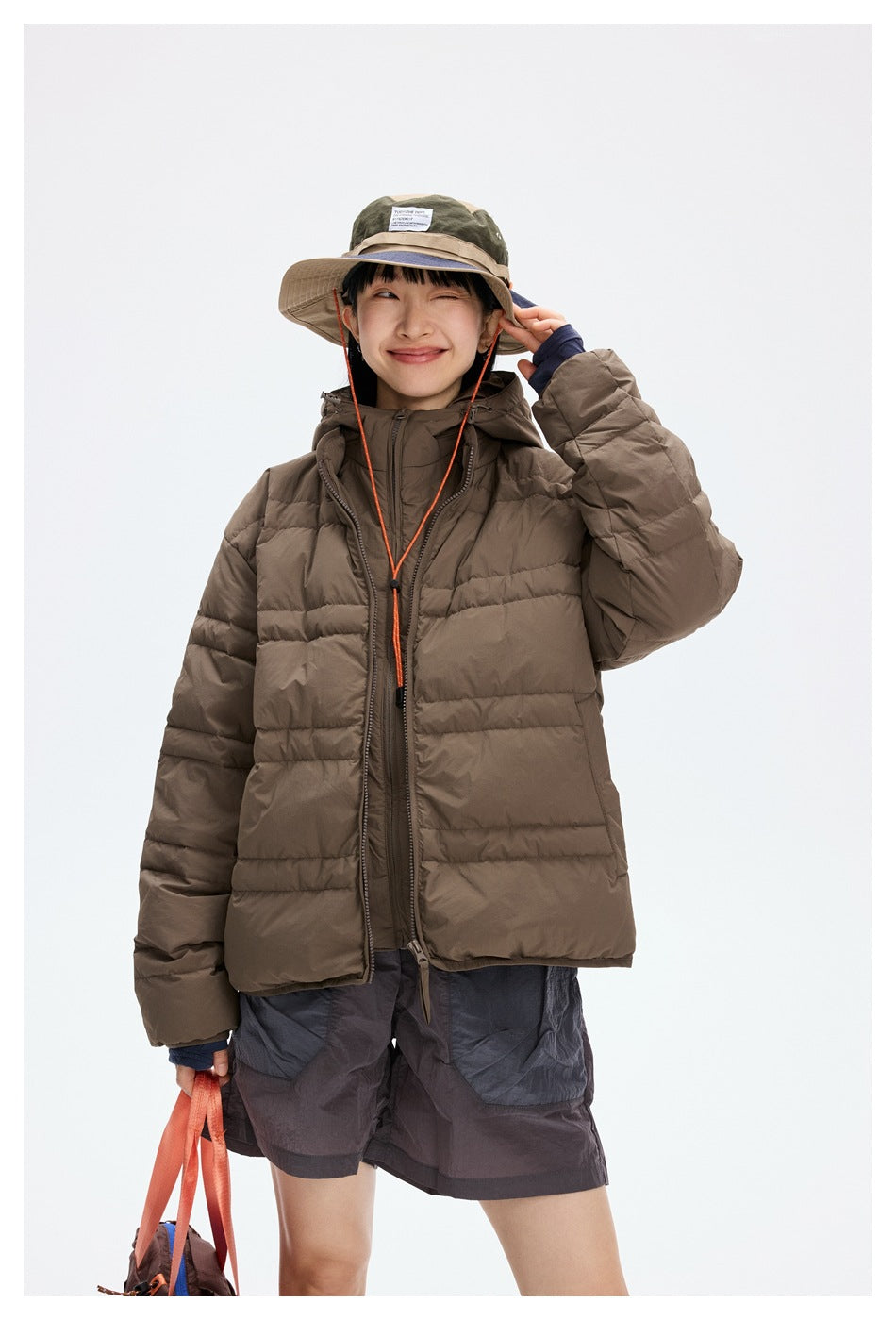 Model in a brown hooded puffer jacket and gray shorts, shown from the front with one hand at her hat.