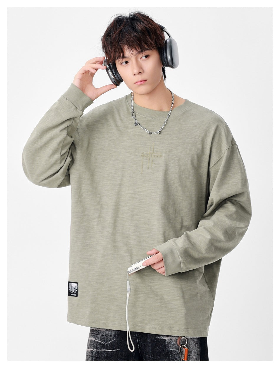 Model wearing a sage-gray oversized long-sleeve crewneck shirt with subtle chest embroidery and a hem patch.