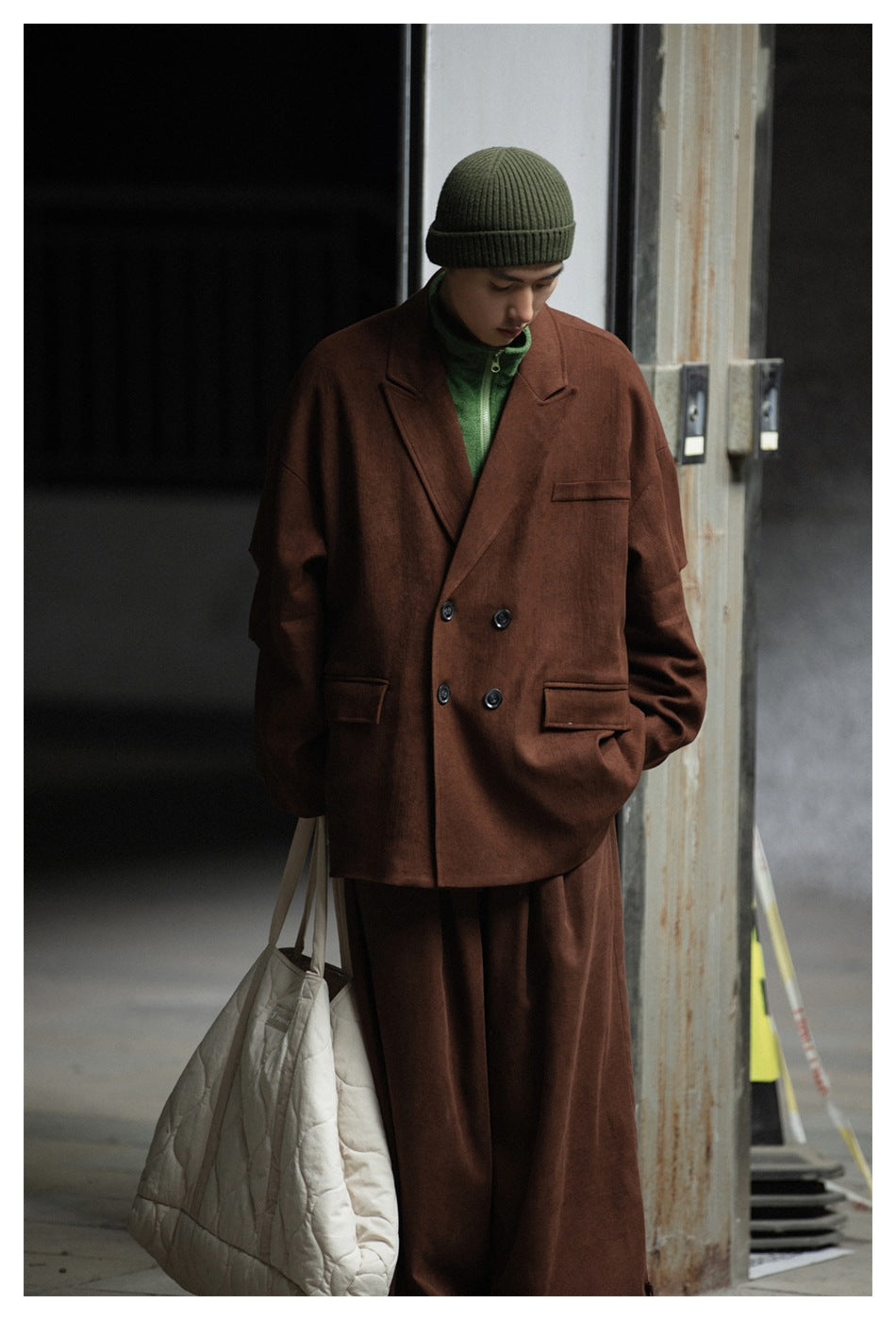 Person wearing a brown double-breasted jacket over a green zip top with a green beanie and a quilted tote bag