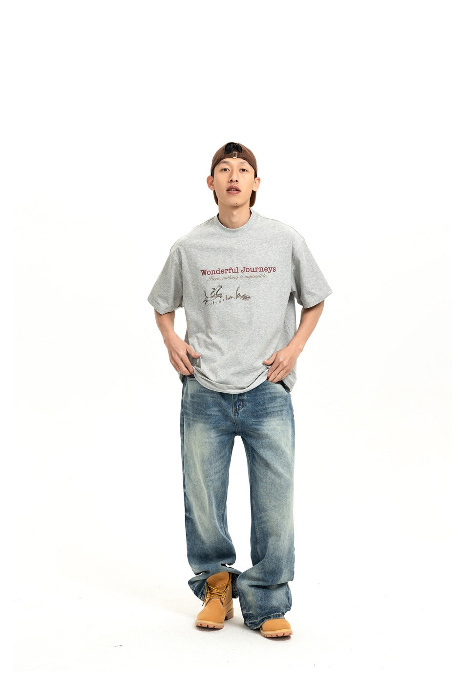 Model wearing the gray Wonderful Journeys T-shirt with faded blue wide-leg jeans, a brown cap and tan boots, front view