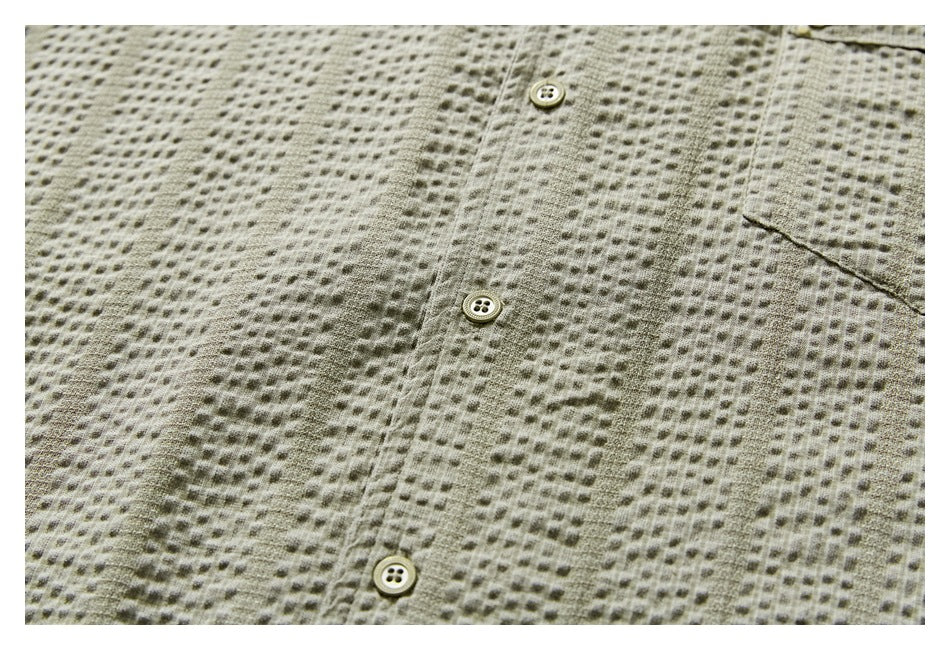 Close-up of a light olive shirt showing its textured fabric, button placket and edge of a chest pocket.