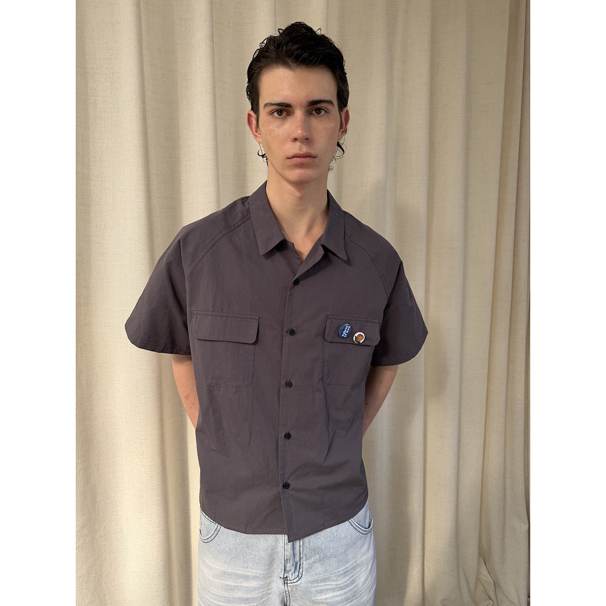 Model wearing a charcoal short-sleeve button-up shirt with two flap pockets and light-wash denim shorts.