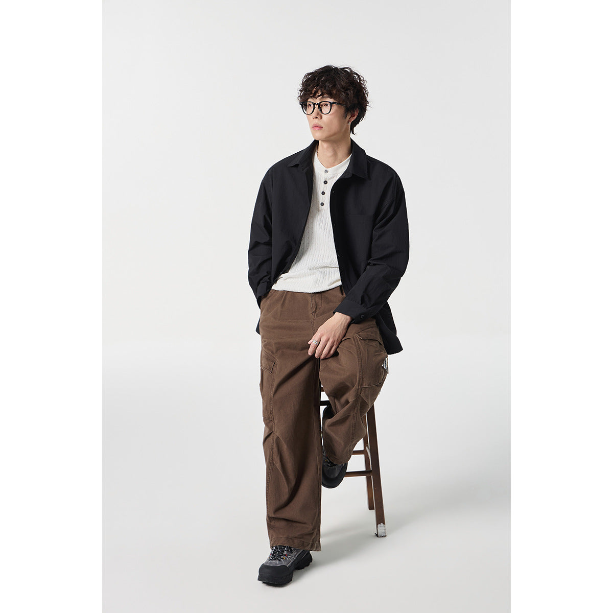 Model wearing a black button-up shirt over a white henley and loose brown cargo pants, seated on a stool.
